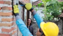 Petugas PLN berhelm kuning memasang meteran listrik prabayar di dinding bata rumah warga