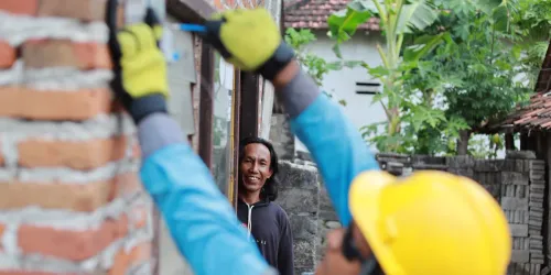 Petugas PLN berhelm kuning memasang meteran listrik prabayar di dinding bata rumah warga